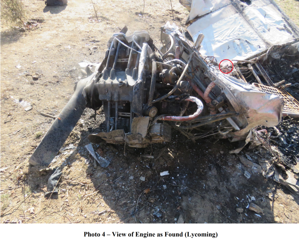 A burned and damaged Lycoming aircraft engine lying on the ground with exposed components, melted wiring, and a broken propeller, surrounded by debris from the crash.