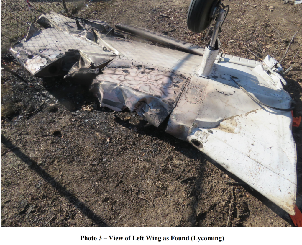 A heavily damaged and partially burned left wing from the crashed aircraft, lying on the ground near a fence, with the landing gear still attached.