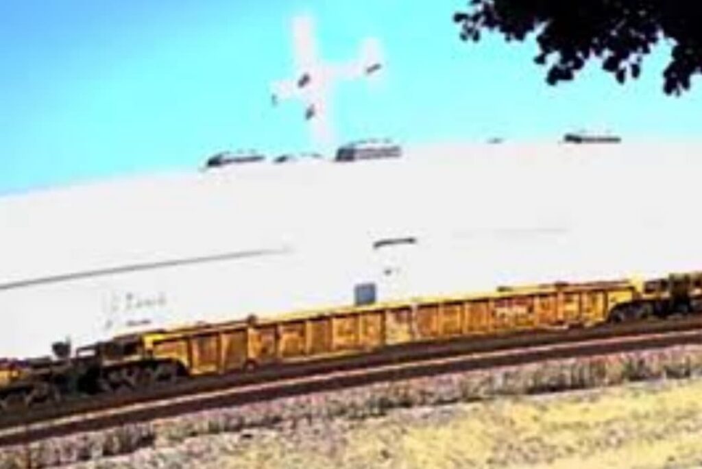 A low-quality image captures an aircraft in a near-vertical nose-down position just above a large white industrial building. The aircraft's wings are visible, and it appears to be in an uncontrolled descent. A yellow freight train is passing in the foreground on a curved track. The sky is bright blue, and part of a tree is visible in the upper right corner.