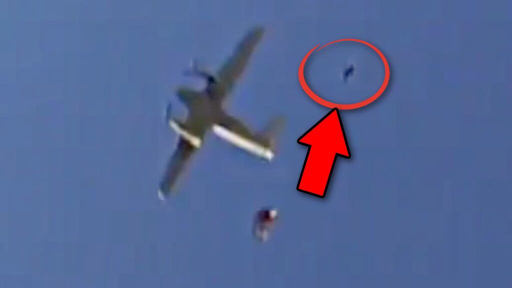 A low-resolution image of an aircraft in flight with two skydivers falling away from it. One skydiver is clearly visible, while another is highlighted with a red circle and arrow, indicating their position in the sky. The background is a clear blue sky.