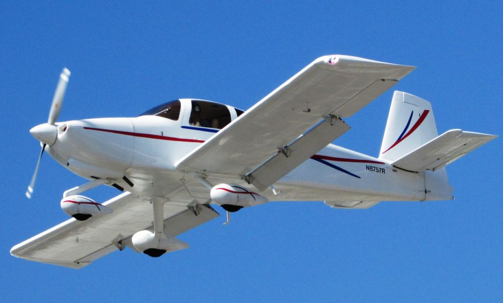 A white, low-wing, experimental Van’s Aircraft RV-10 with red and blue accents is flying against a clear blue sky. The aircraft's registration number, N8757R, is visible on the fuselage near the tail. The landing gear appears to be extended, and the propeller is spinning.