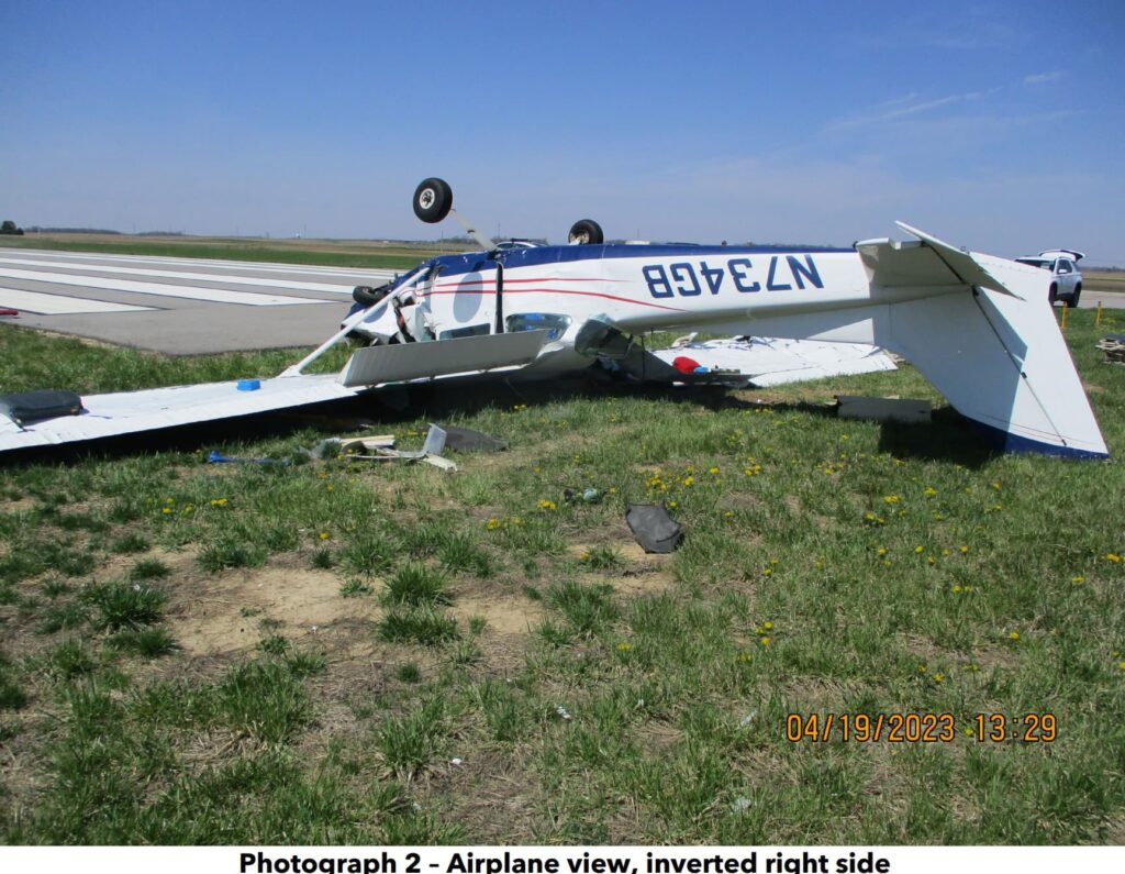 A wrecked Cessna 172 (registration N734GB) resting upside-down in a grassy field near a paved surface. The fuselage is crushed, with significant damage to the nose section. The left wing appears torn off and lies nearby. Various debris, including aircraft components and personal belongings, are scattered around. A rural landscape with trees and farm structures is visible in the background.