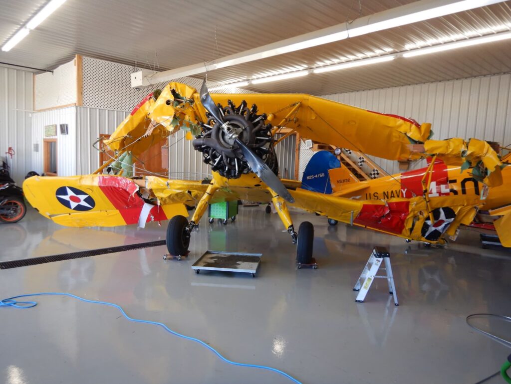 A severely damaged yellow Boeing Stearman biplane inside a hangar. The upper and lower wings are crumpled, and the propeller is bent. The fuselage displays "U.S. Navy" markings, and parts of the aircraft are wrapped in green straps for support.