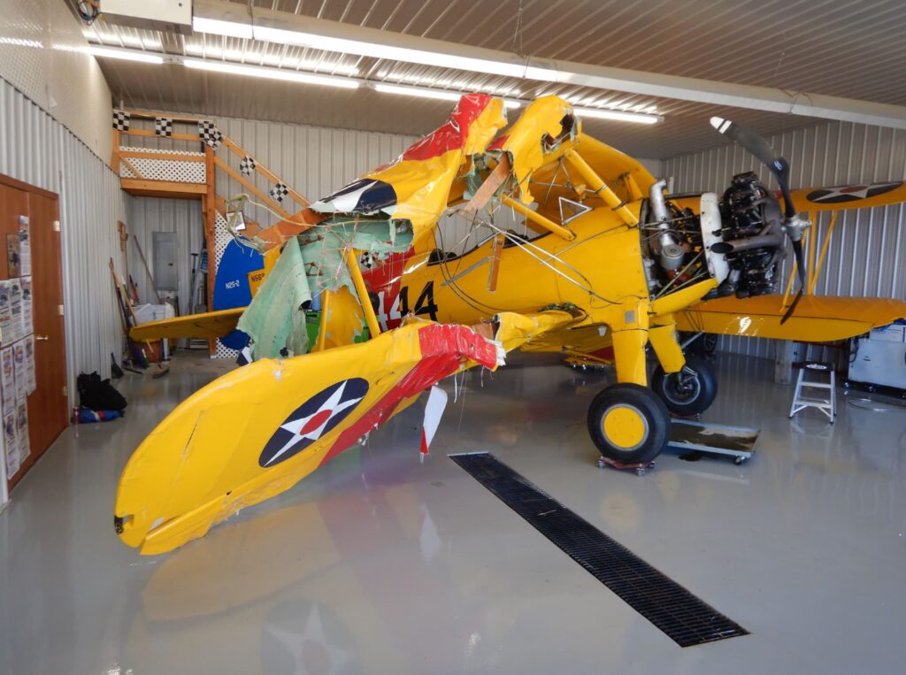 A side view of the wrecked Stearman biplane inside the hangar. The left wing is crushed and torn, revealing internal structures. The radial engine is exposed, and the aircraft's nose is slightly tilted downward.