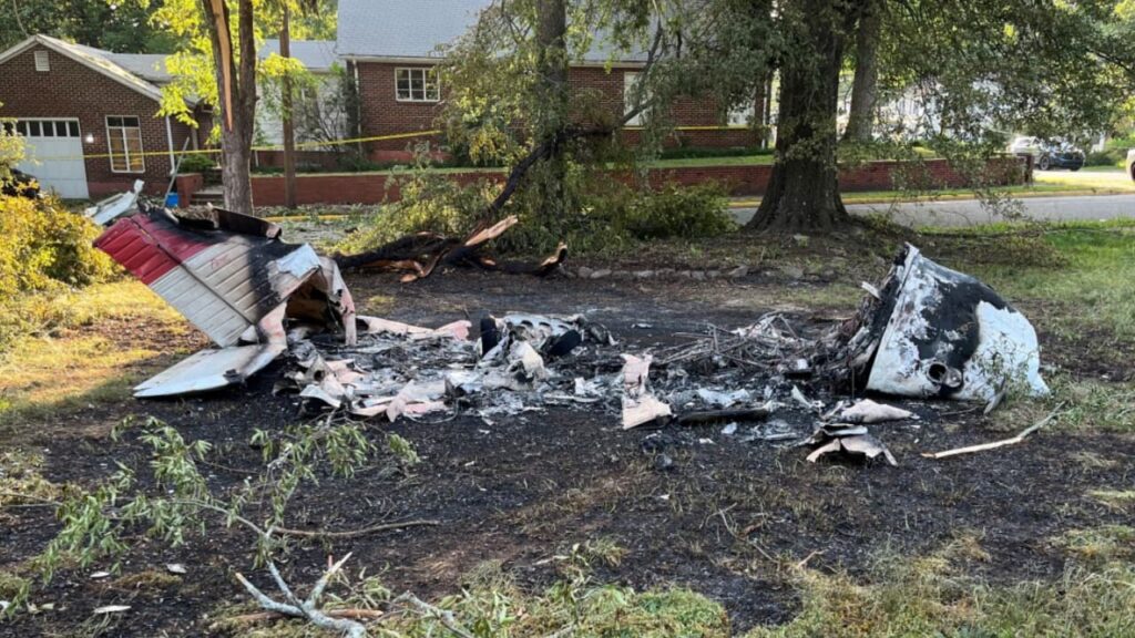 Burned-out wreckage of a Piper PA-28-180 aircraft in a vacant lot near a tree-lined street. The remains are scattered, with the fuselage almost entirely destroyed by fire.