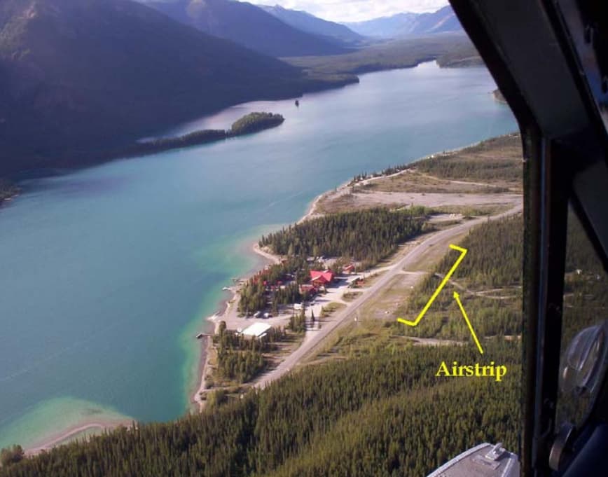 An aerial view of the remote airstrip at Muncho Lake, British Columbia, surrounded by dense forests and a large body of water. The small gravel airstrip is highlighted with yellow markings, showing its short length and proximity to the lodge and highway