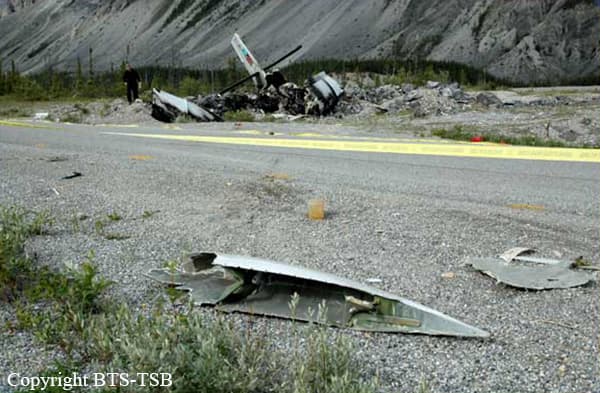 "Wreckage of a De Havilland DHC-6 Twin Otter scattered near a remote highway after a failed takeoff. Debris is visible in the foreground, while the charred remains of the aircraft rest against a rocky embankment. A person stands in the distance near the crash site, and yellow caution tape marks the area. Rugged mountains and trees are in the background