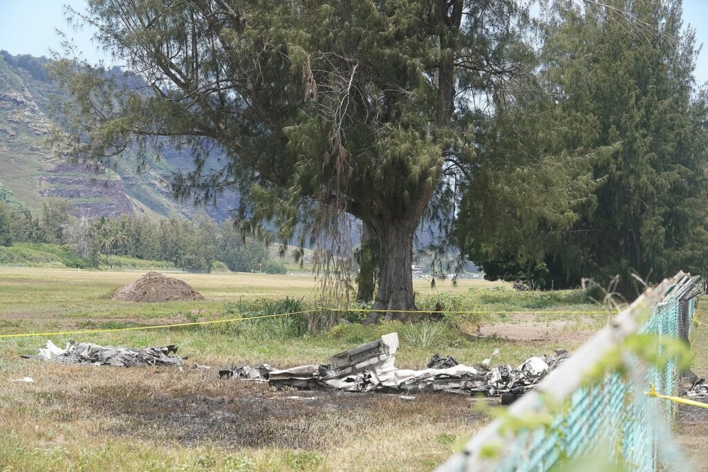 Scattered aircraft wreckage lies on the grass near a tree at Dillingham Airfield, Hawaii. A burned section of debris is surrounded by yellow caution tape, with a distant mountain range and greenery in the background.