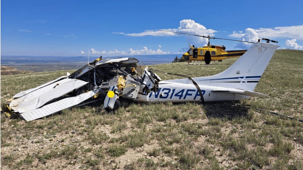 "Front view of a severely damaged Cessna 182S aircraft wreckage resting on a grassy field in mountainous terrain. The aircraft's fuselage is crushed, and the wings are heavily crumpled. A yellow rescue helicopter is visible in the background under a bright blue sky with scattered clouds."