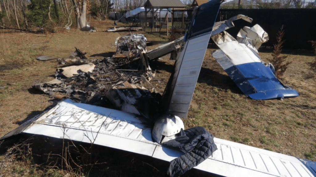 Wreckage of a Piper PA-28-161 aircraft after a crash, showing charred and twisted metal remains, burned fuselage, and broken wings in an open area with trees and a playground structure in the background.