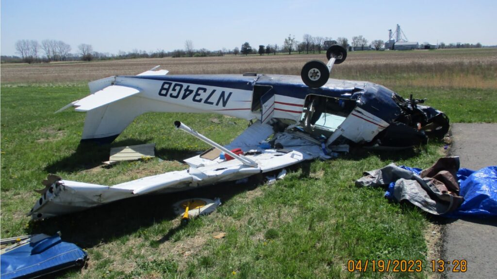 An overturned Cessna 172 aircraft (registration N734GB) lying inverted on a grassy area near the edge of a runway. The aircraft’s right wing is severely damaged, and debris is scattered around. The landing gear is fully extended, facing upward. The sky is clear, and the airport environment is visible in the background.