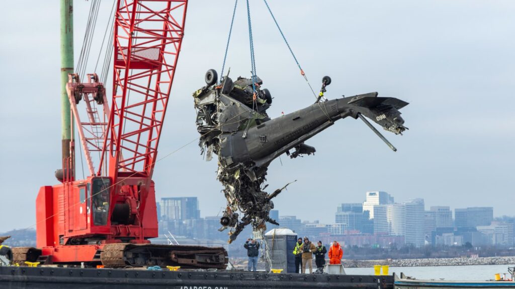 A large crane lifts the wreckage of a heavily damaged UH-60 Blackhawk military helicopter from the water onto a barge, with emergency personnel and onlookers nearby. A city skyline is visible in the background.