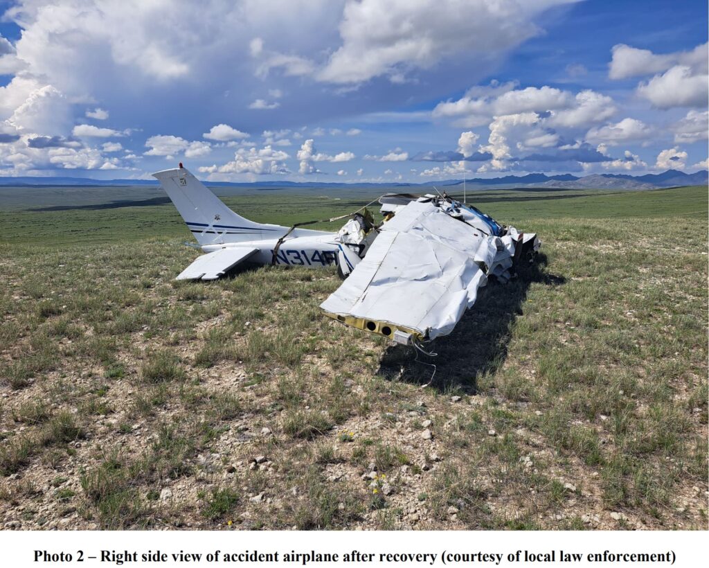 Right side view of a crashed Cessna 182S airplane in a remote, open field with rolling hills in the background. The fuselage is extensively damaged, with the right wing bent and crumpled. The tail section remains intact, displaying the aircraft registration number N314FR. The sky is partly cloudy with clear visibility.