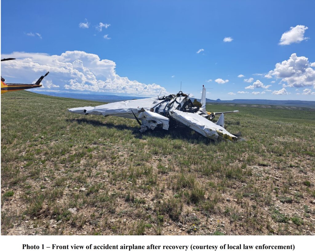 Left side view of a wrecked Cessna 182S airplane resting on uneven grassy terrain. The fuselage is crushed, with the cockpit severely deformed. The left wing is crumpled and partially detached. A yellow rescue helicopter is parked nearby, with rolling hills and a distant horizon under a partly cloudy sky.