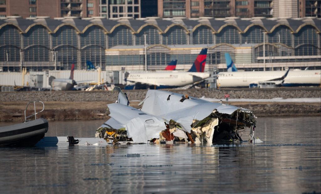 Wreckage of the American Airlines Flight 5342 aircraft partially submerged in the Potomac River near the Ronald Reagan National Airport in Washington DC, with a rescue boat nearby. In the background, Delta Air Lines aircraft are parked at a terminal.