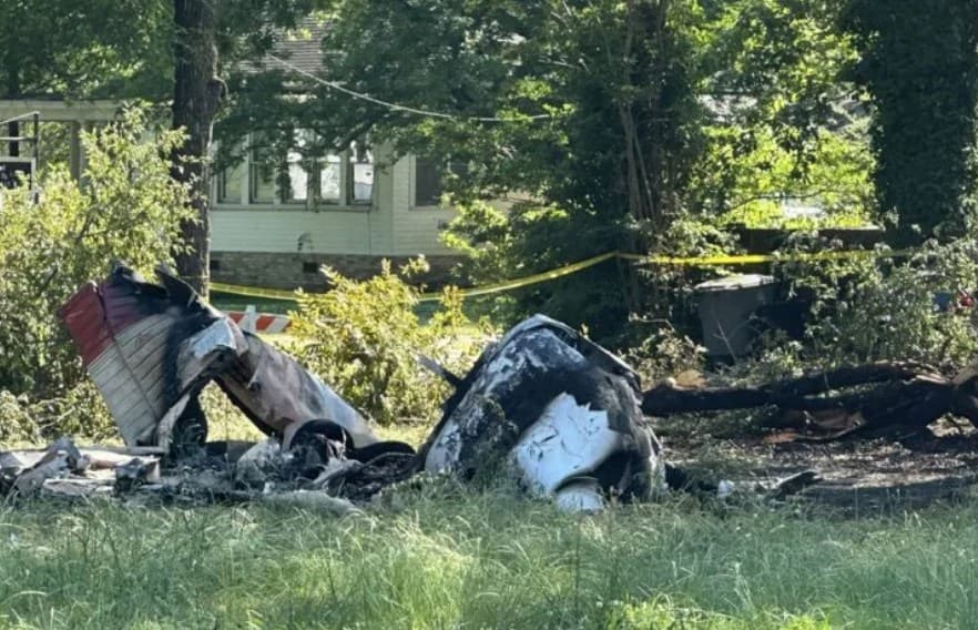 Charred remains of a Piper PA-28-180 aircraft in a grassy residential area, with a house and yellow caution tape in the background. The aircraft is severely burned, showing extensive fire damage.