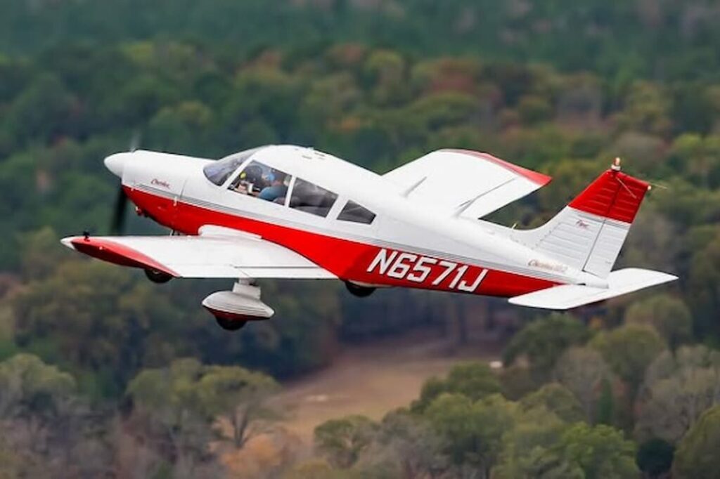 A red and white Piper PA-28-180 with registration N6571J flying over a forested area. The aircraft is in level flight, with a clear canopy showing the pilot and passenger inside.