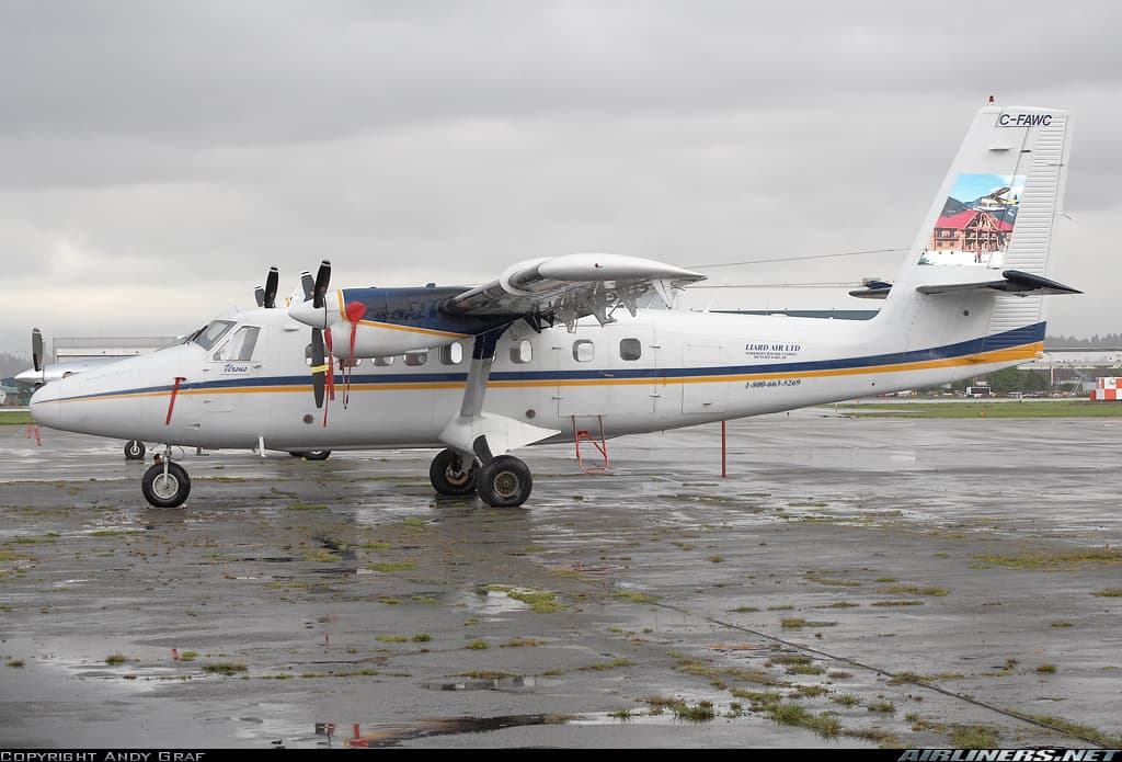 A De Havilland DHC-6 Twin Otter, registered C-FAWC, parked on a wet tarmac under an overcast sky. The aircraft belongs to Liard Air Limited and features a colorful lodge-themed tail design. Red protective covers are placed on the propellers and engine inlets.