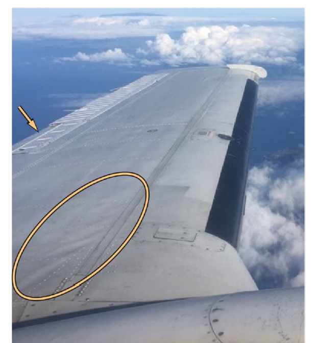 A close-up view of an aircraft wing taken mid-flight, showing wrinkled skin near the leading edge. A yellow circle highlights the damage, and an arrow points to another area of concern. Below, the ocean and clouds are visible.