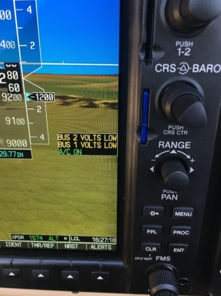 A close-up photograph of an aircraft avionics display showing flight information and warning messages. The screen displays a terrain view, altitude, and voltage alerts indicating "BUS 2 VOLTS LOW" and "BUS 1 VOLTS LOW." The text "A/C ON" is also visible, along with various navigation and system control buttons on the right side of the panel