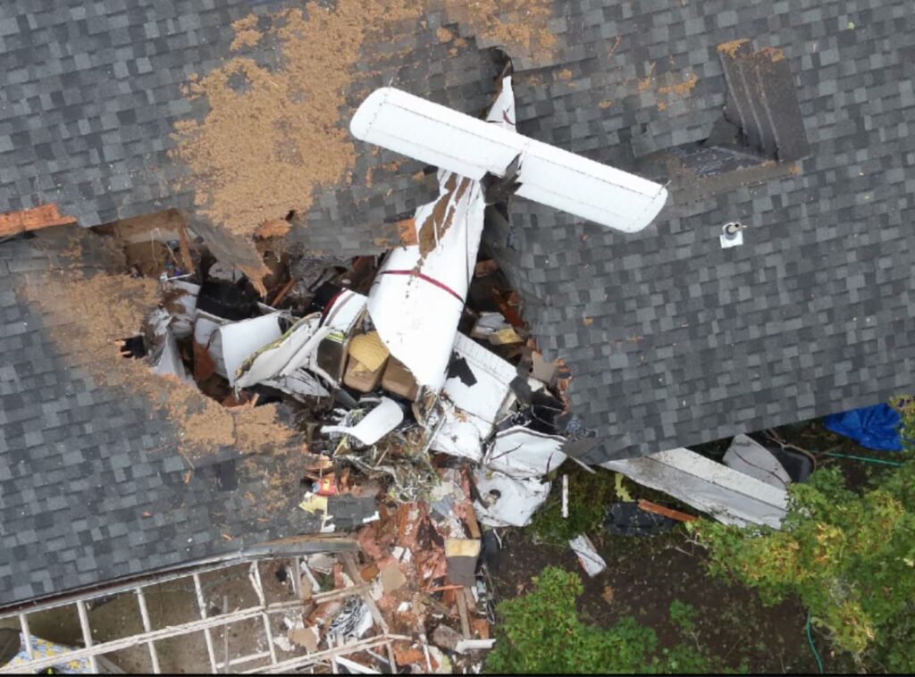 Overhead view of a Piper PA-44 Seminole aircraft crashed through the roof of a single-story house, with debris and broken roof shingles surrounding the fuselage and tail.