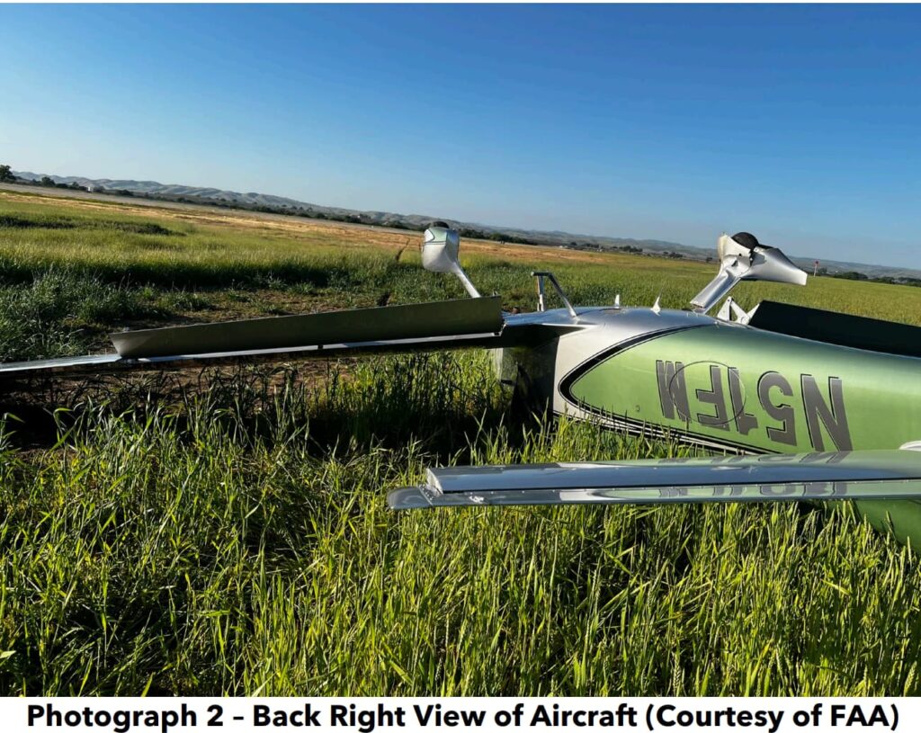 An overturned Cirrus SR22T aircraft resting in a grassy field, showing significant damage to the wings and fuselage. The aircraft’s registration number, "N51FM," is visible upside down on the fuselage. The surrounding landscape includes open fields and distant hills under a clear blue sky.