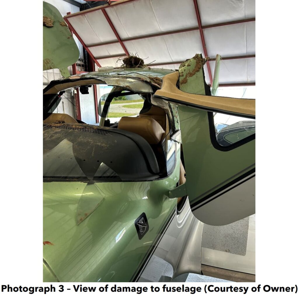 A close-up view of the damaged fuselage of a Cirrus SR22T inside a hangar. The aircraft's roof is severely crushed, with dirt and debris lodged in the damaged section. The tan leather interior is visible through the damaged cockpit area, and part of the aircraft’s gull-wing door is bent and open.