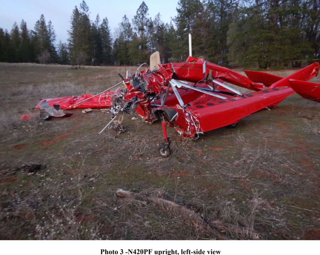 The wreckage of a red aircraft sits in an open field, its structure mangled and wings bent. The landing gear is partially intact, but the front section is destroyed. Surrounding trees and a dirt clearing provide context to the crash site.