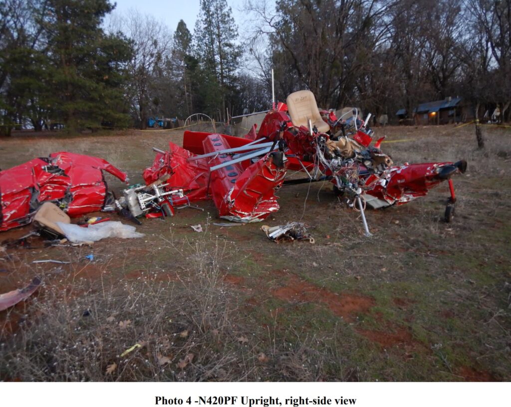 A heavily damaged red aircraft rests on uneven terrain, its fuselage crushed and wings severely crumpled. The exposed cockpit reveals a single beige seat with tangled wiring, and the front structure appears torn apart. Trees and a small house are visible in the background.