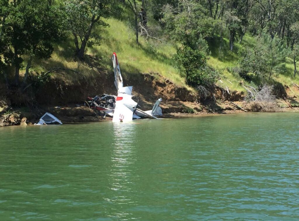 A partially destroyed ICON A5 aircraft sits at the edge of a lake, with its fuselage torn apart. The aircraft’s tail remains upright while debris is scattered along the shoreline. Trees and a steep hillside surround the crash site.