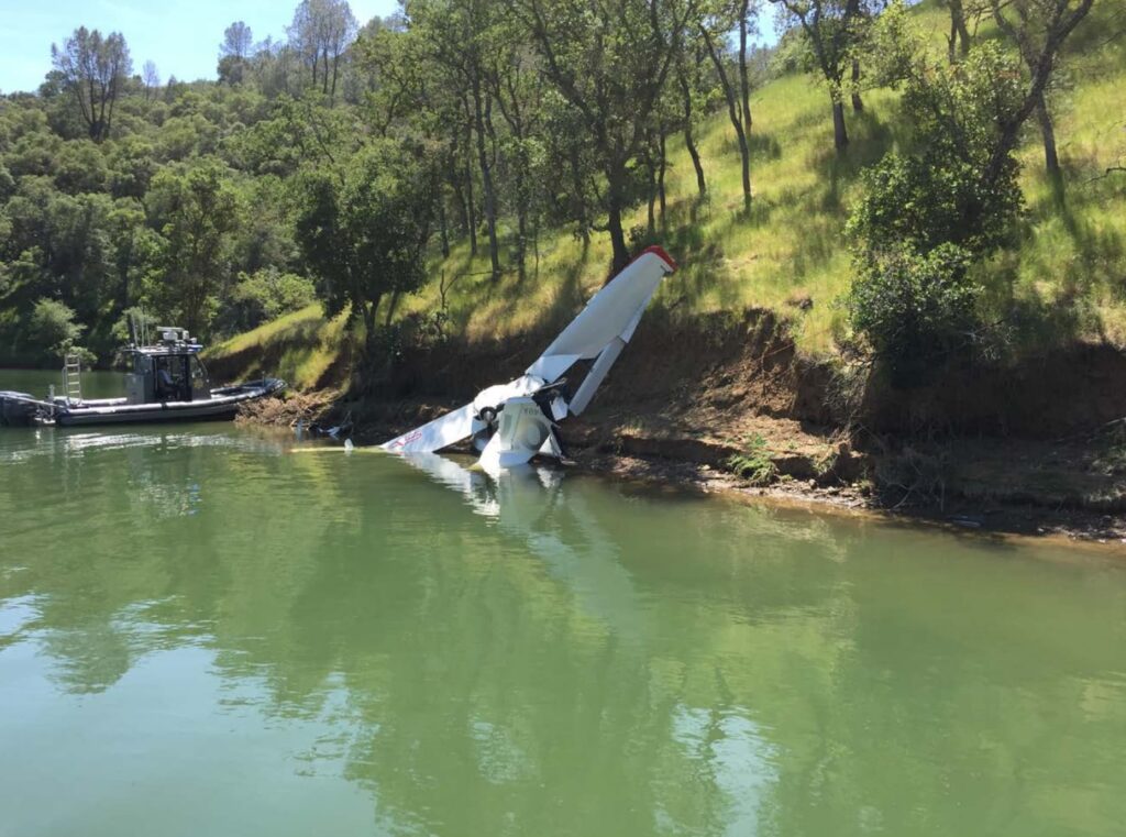 The wreckage of an ICON A5 lies partially submerged along the shoreline of Lake Berryessa. A police boat is nearby, and the aircraft’s tail stands upright against a grassy hillside.