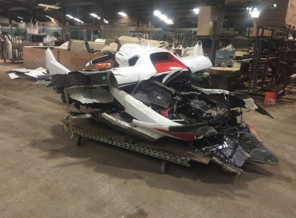 A severely damaged ICON A5 aircraft rests on a wheeled platform inside a storage facility. The fuselage is crushed, with shattered components and tangled wiring visible. Surrounding the wreckage are shelves with aviation parts and equipment.