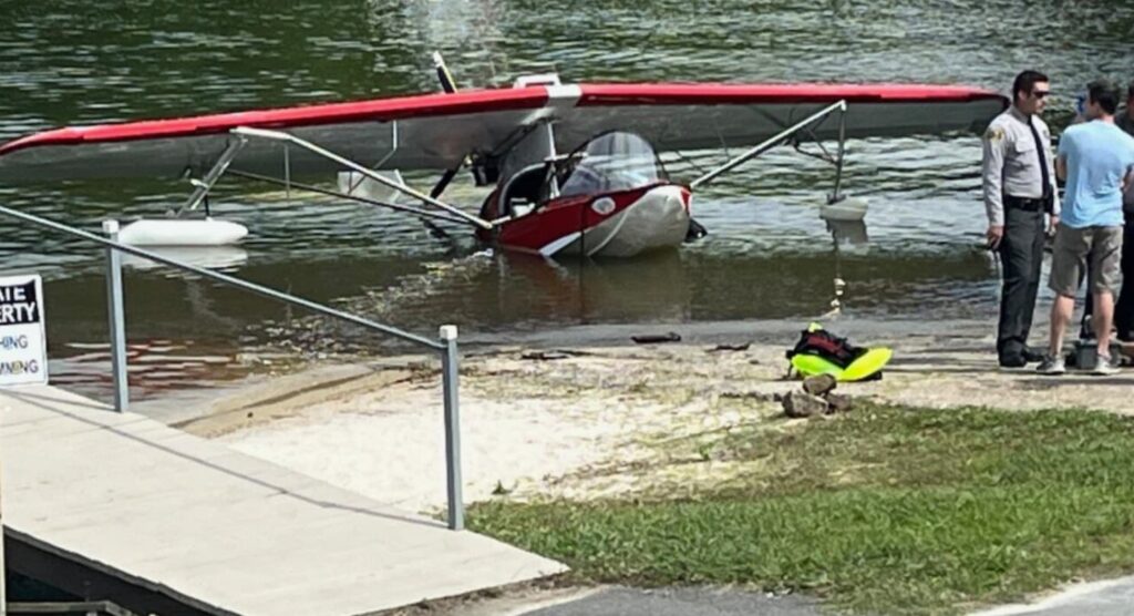 Partially submerged Searey aircraft at the edge of a lake as a law enforcement officer and bystanders discuss the incident nearby.