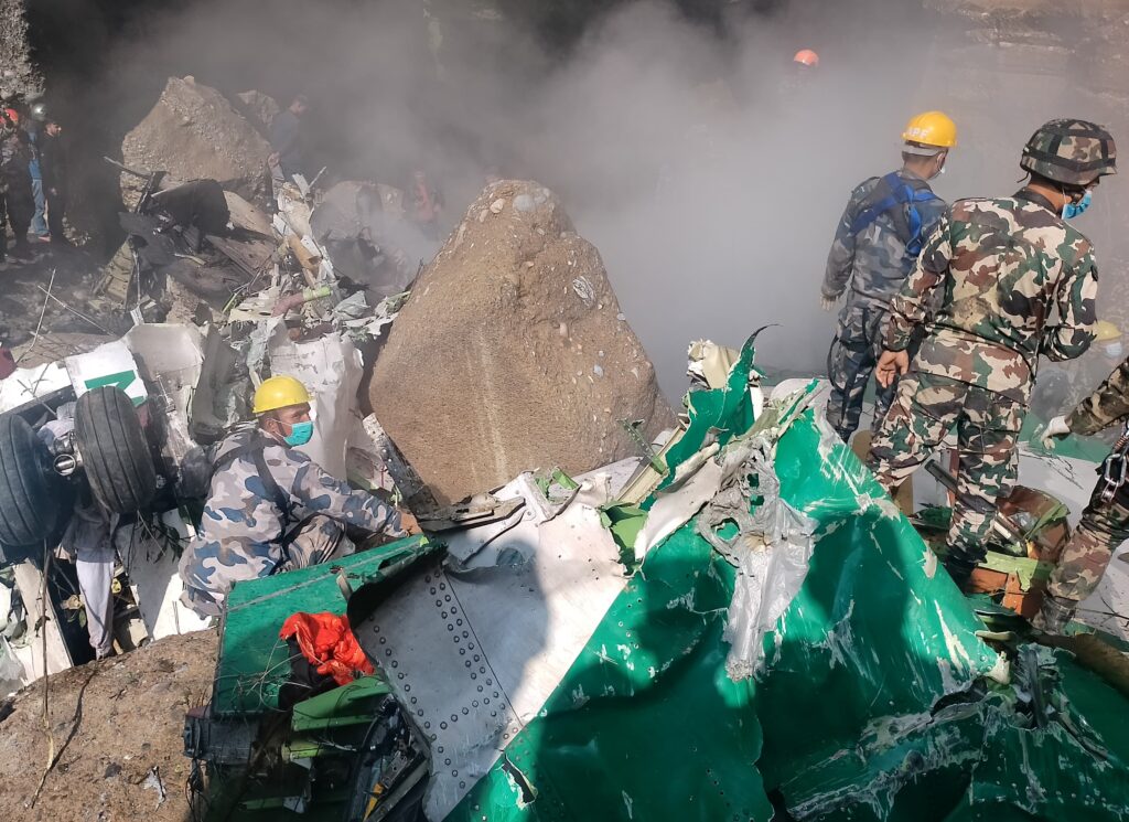 Rescue workers and military personnel search through the wreckage of Yeti Airlines Flight 691, which crashed near Pokhara, Nepal. The image shows debris from the ATR 72 aircraft, including twisted metal, a damaged landing gear, and burned fuselage fragments. Thick smoke is rising from the crash site as rescuers in camouflage uniforms and safety helmets sift through the wreckage.