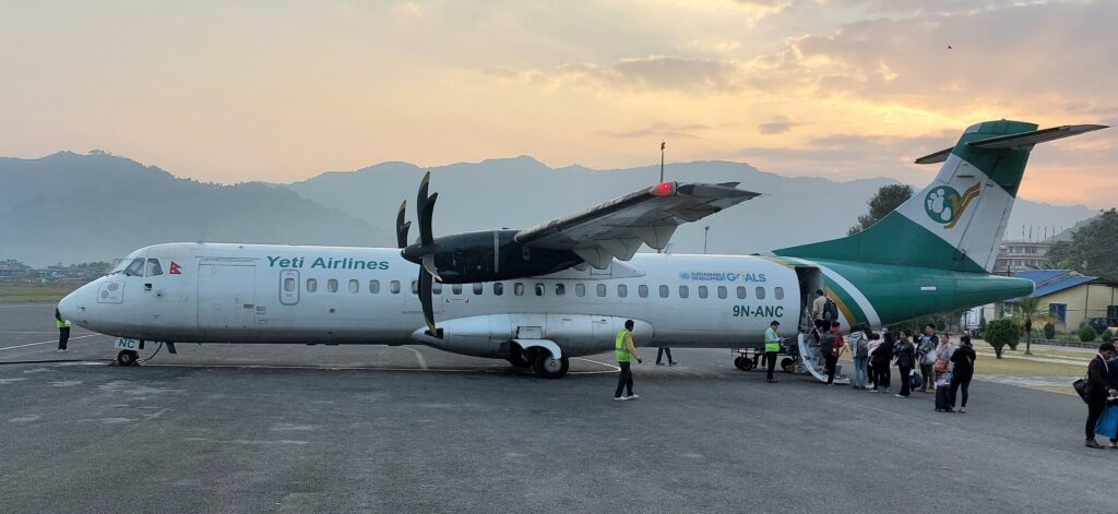  Yeti Airlines ATR 72-212A (registration 9N-ANC) parked at an airport with passengers boarding via the rear stairs. The aircraft is painted in green and white, displaying the Yeti Airlines logo on the tail. Ground crew in high-visibility vests are assisting with boarding. The backdrop features a mountainous landscape under a sunset sky.