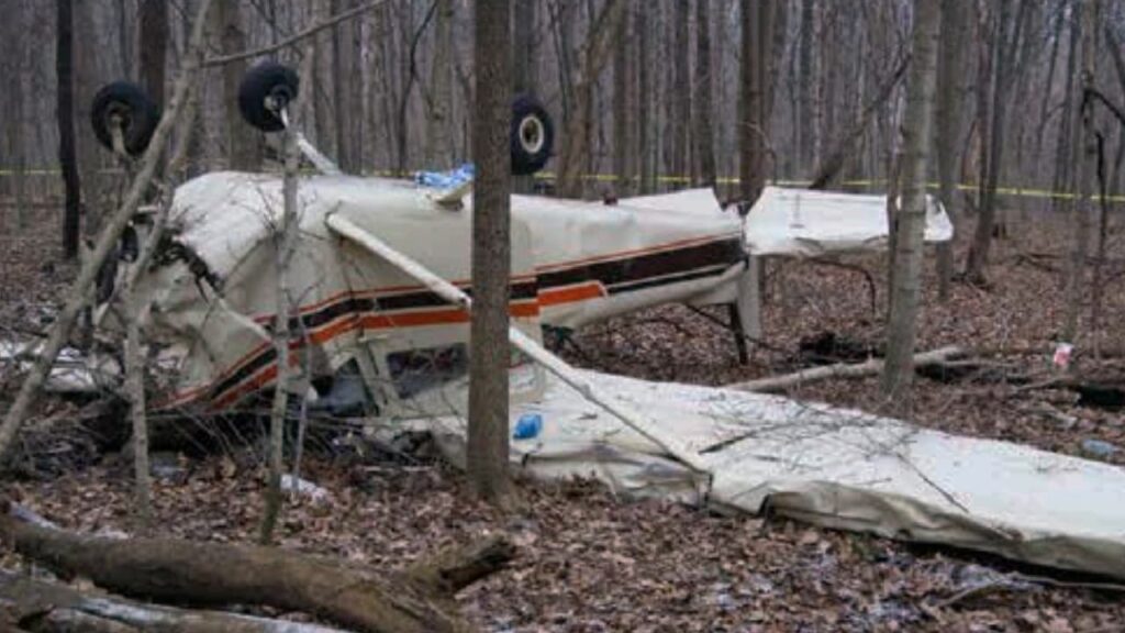 Wreckage of a Cessna 172F aircraft lying inverted in a wooded area after a crash. The fuselage is severely damaged, with the wings partially detached and the landing gear visible above the overturned aircraft. Surrounding trees and fallen debris indicate the aircraft impacted terrain in a forested location. Yellow caution tape is seen in the background, marking the crash site.