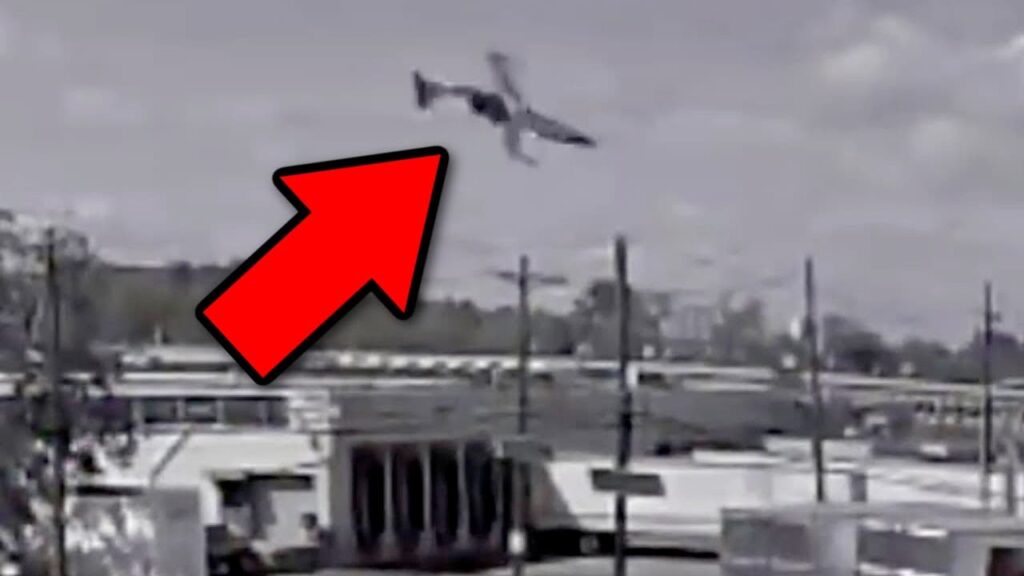 A black-and-white security camera still frame showing an aircraft in a steep nose-down, nearly inverted position just before impact. A large red arrow points to the aircraft, emphasizing the dramatic moment. The background includes buildings, power lines, and an overcast sky, indicating an urban or industrial area.