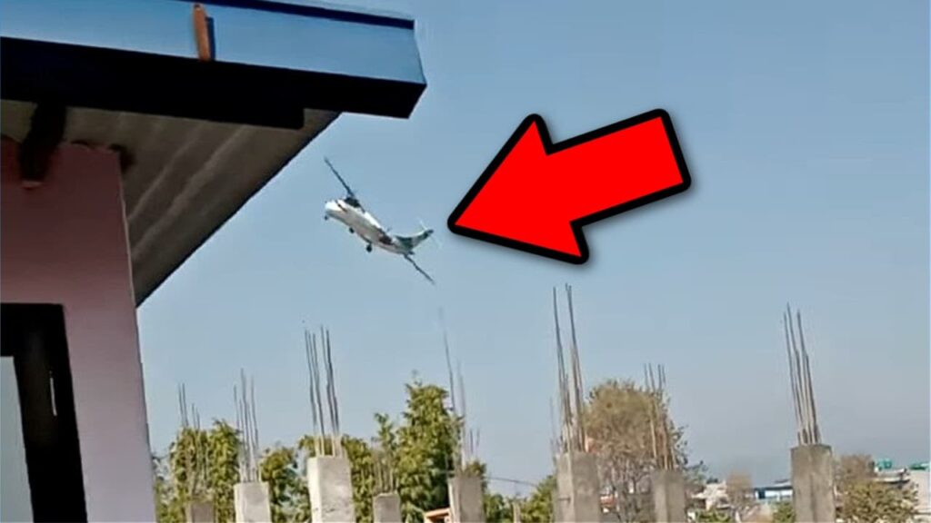 A still image of Yeti Airlines Flight 691, an ATR 72, moments before crashing near Pokhara, Nepal. The aircraft is seen in a steep left bank with its nose pointing downward. A large red arrow highlights the plane. The foreground shows part of a building with exposed construction rods, and the background features a clear blue sky.