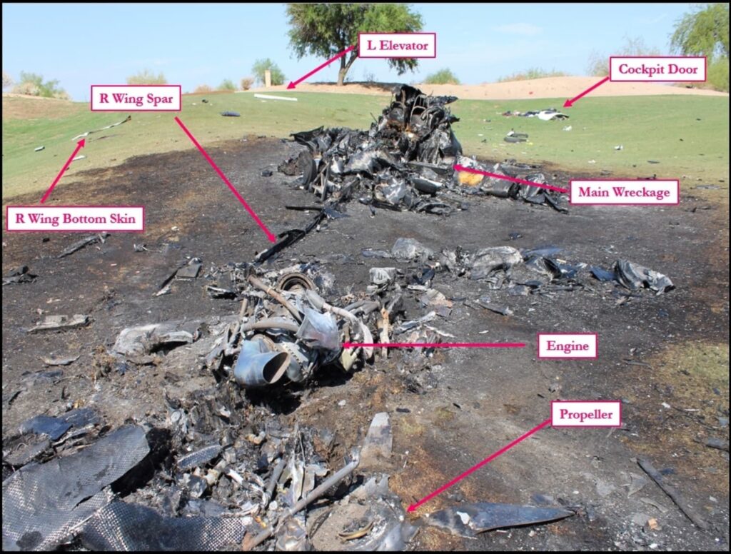 A photograph of a burnt aircraft crash site with various wreckage components labeled. The charred remains of the aircraft are scattered across a grassy area with blackened ground. Labels indicate different parts of the wreckage, including the "Engine," "Propeller," "Main Wreckage," "Cockpit Door," "L Elevator," "R Wing Spar," and "R Wing Bottom Skin." The aircraft is heavily damaged, with only fragmented and burnt remains visible. The background shows a green grassy hill with debris spread across the area.