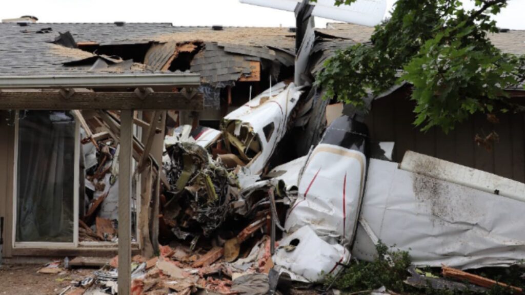 Side view of the heavily damaged Piper PA-44 Seminole aircraft lodged into the side of a house, with crumpled wings and shattered walls visible around the wreckage.