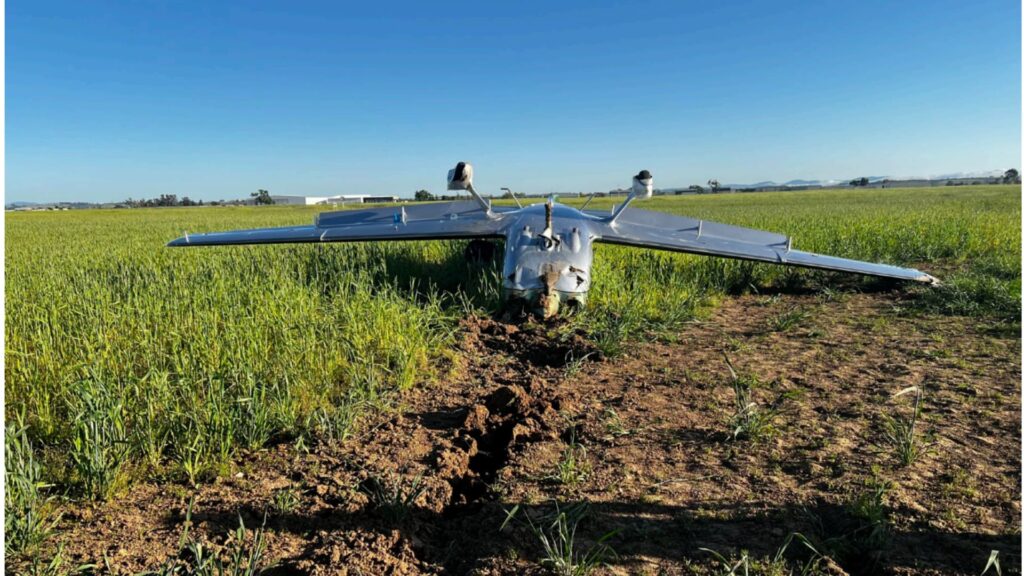 An overturned Cirrus SR22T aircraft lying in a grassy field, viewed from the rear. The aircraft's wings are intact, but the fuselage is heavily damaged. A trail of disturbed soil leads to the wreckage, indicating the aircraft’s path before coming to rest. The landing gear is exposed, and the clear blue sky contrasts with the accident scene.