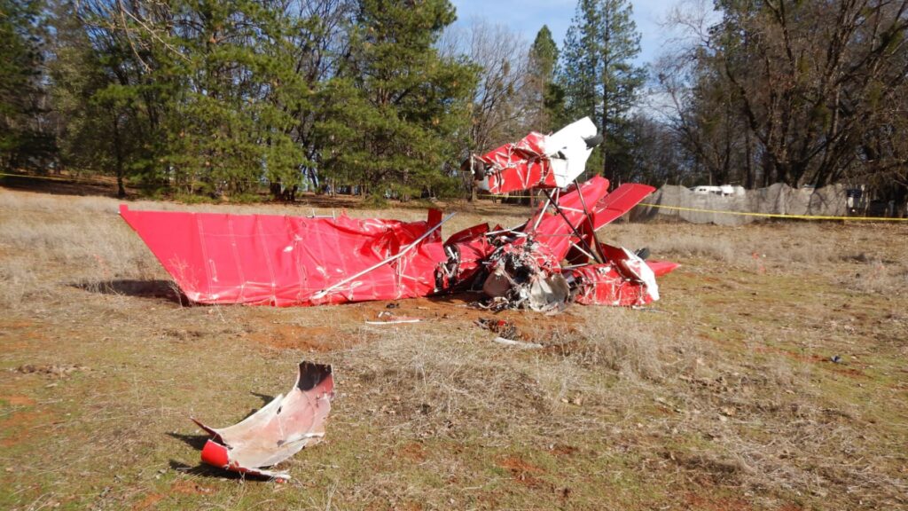 A red aircraft's wreckage is scattered across a rural field, with the front section almost unrecognizable due to extensive impact damage. A detached piece of the aircraft lies in the foreground, while yellow caution tape marks the perimeter of the crash site. Trees and a residential area are visible beyond the wreckage.