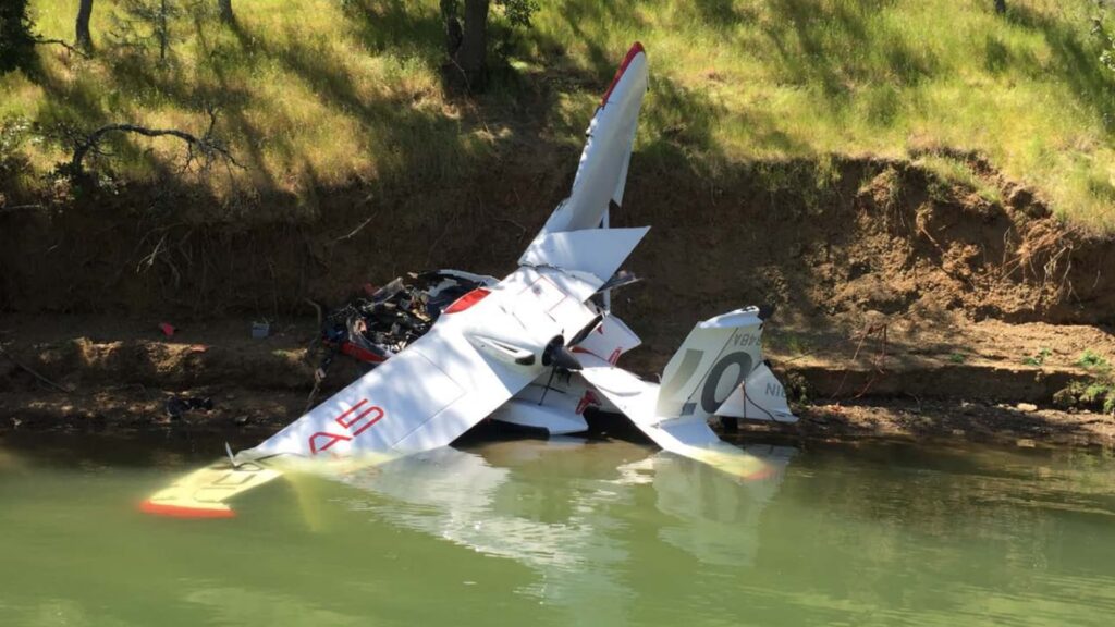 The ICON A5's crushed fuselage and broken wings rest against a dirt embankment, with one wing partially submerged in the lake. The aircraft's registration number is partially visible, and wreckage is strewn along the shoreline.