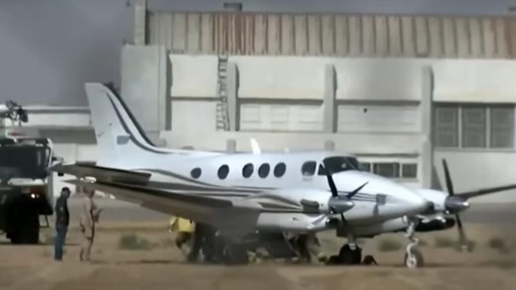 A white twin-engine turboprop aircraft, a Raytheon C90A King Air, parked on a dirt surface near an airport building. Several emergency responders and personnel are gathered around the plane, with a fire truck visible nearby. The scene appears to show an emergency response following an incident involving the aircraft.