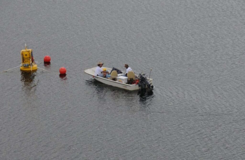 Overhead view of two researchers in a small motorboat on a calm lake near a yellow scientific buoy and red marker floats, taken from a low-flying aircraft.