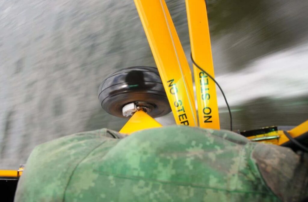 Close-up view from inside a low-flying Piper aircraft showing the yellow landing gear strut and main tire just above the water surface, with visible water spray and part of a passenger’s camo-patterned pants.