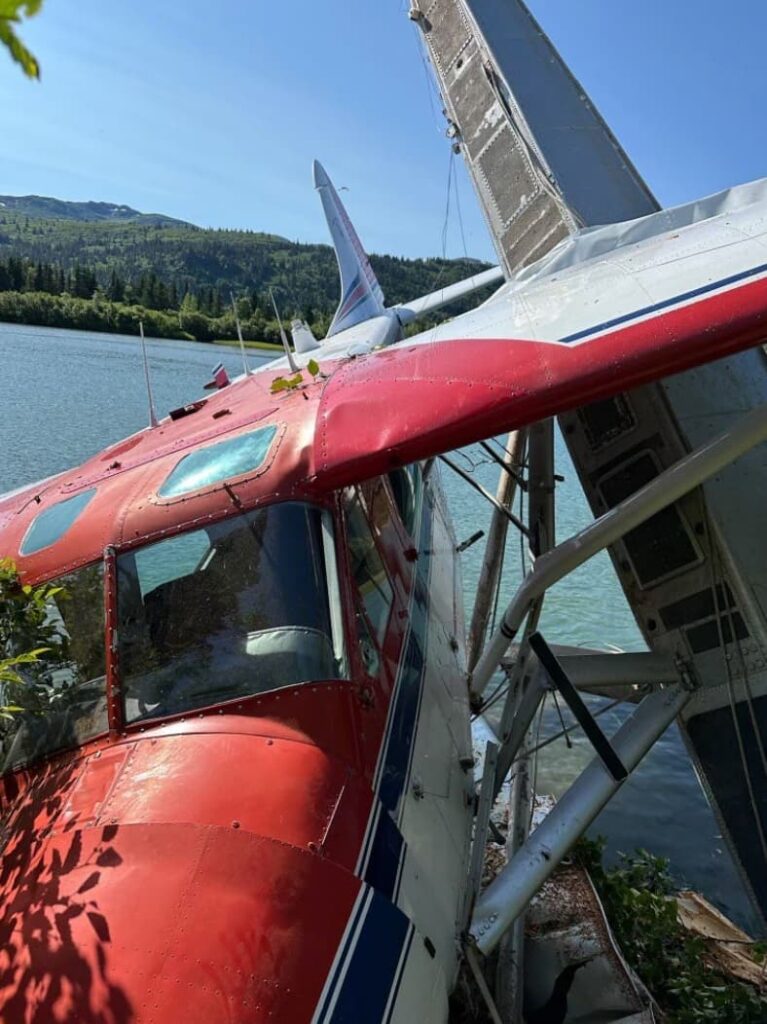 Close-up of DeHavilland Beaver U-6A cockpit and wings after crash, showing bent wing struts and minor foliage entanglement, with mountains and lake in background.