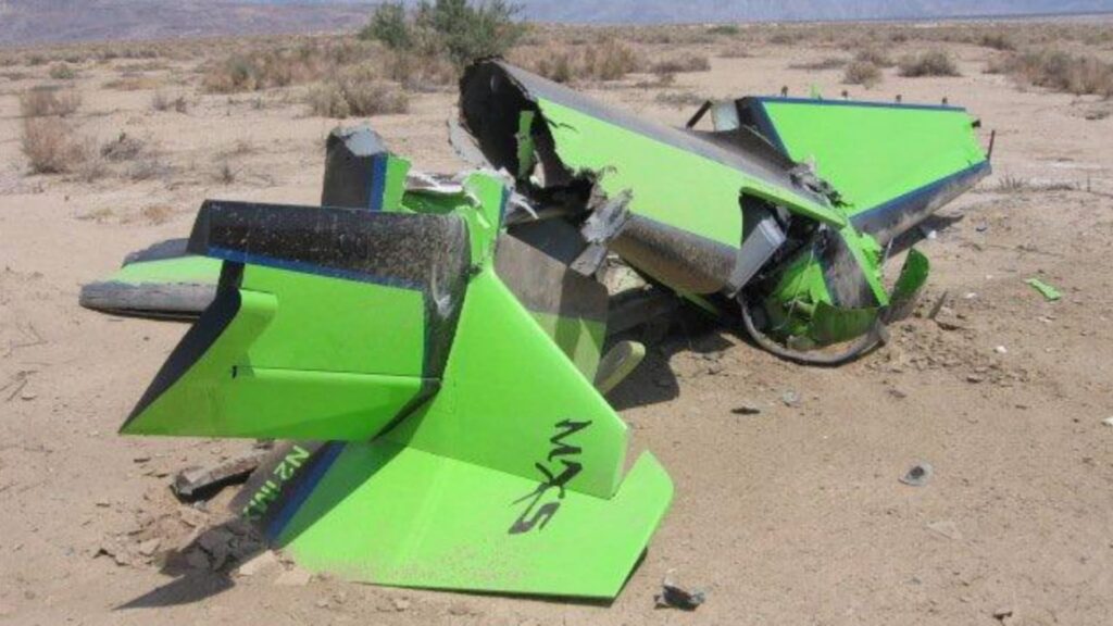 Wreckage of a bright green MXS aerobatic aircraft lying in a desert area, with the tail section heavily damaged and broken off. The aircraft is resting on sandy ground with sparse vegetation in the background. The registration "N21MX" and "MXS" logo are visible on the tail.