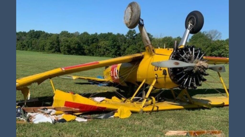 An overturned yellow Boeing Stearman biplane rests on a grassy field with substantial damage. The top wing is crumpled, the propeller is bent, and the aircraft’s tail is broken. One wheel is visibly raised in the air, and scattered debris is visible around the wreckage. Trees line the background under a clear blue sky.