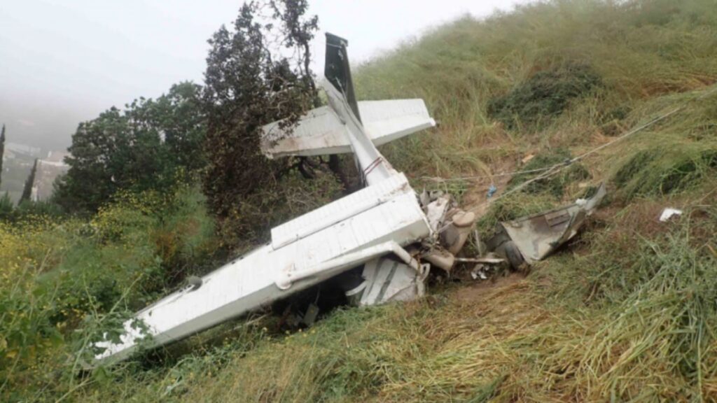Wreckage of a small white aircraft lying crumpled on a grassy hillside, with the tail section partially intact and the fuselage heavily damaged. The aircraft is surrounded by tall grass and vegetation, with fog and trees visible in the background, suggesting a mountainous or hilly terrain.